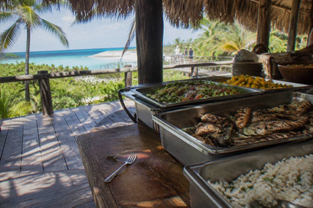 Buffet table on a shaded deck with trays of rice, grilled fish, salad and fruit