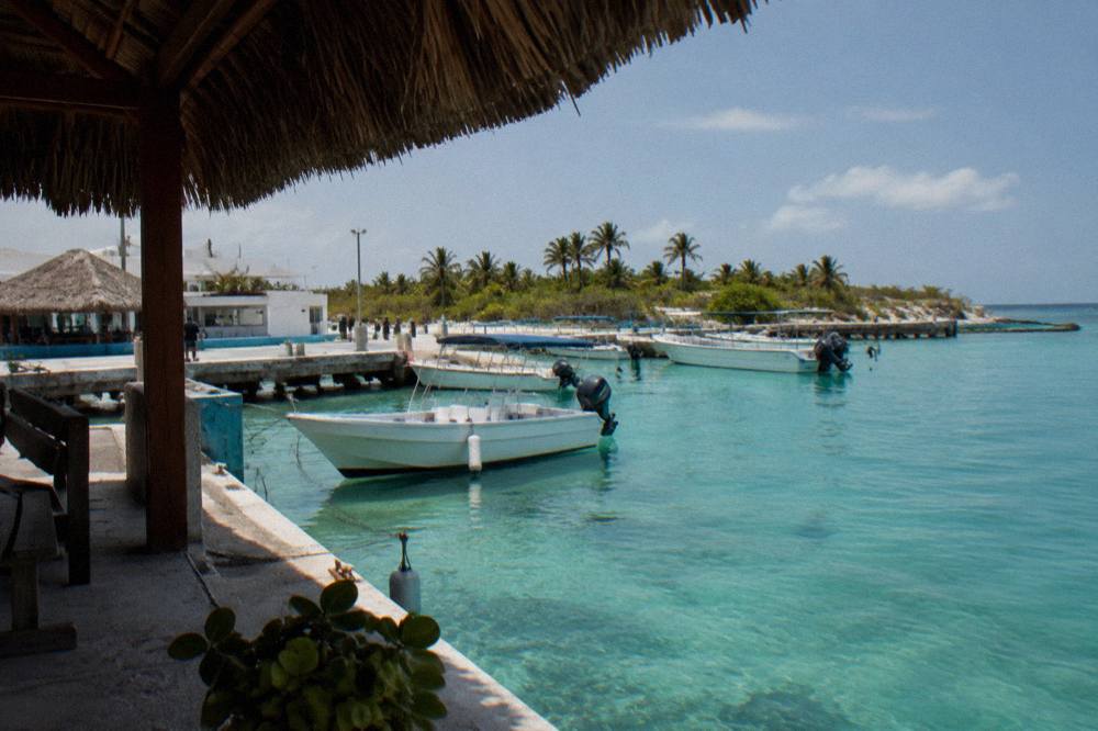 A quiet white sand beach with shallow turquoise water and a few moored boats in the distance