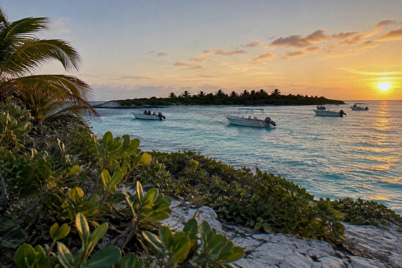 Small boats anchored off a low green island with white sand and calm turquoise water