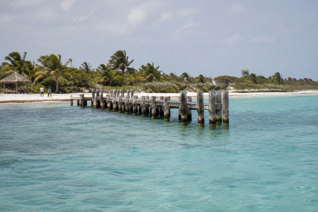A white sandy island shoreline with calm turquoise water and a wooden pier in the distance