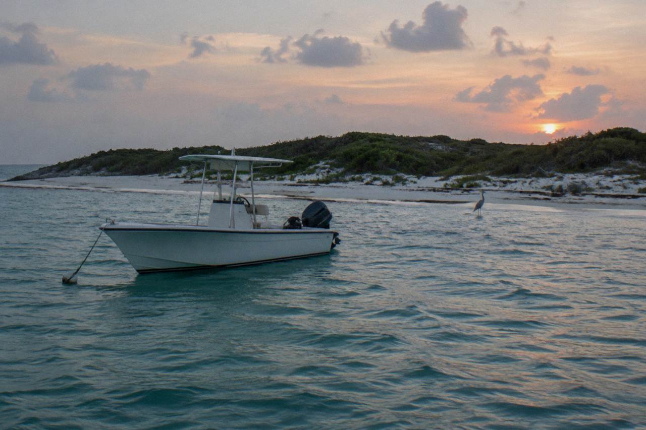 A small white speedboat anchored off a palm-free sandy island shoreline under a wide sky