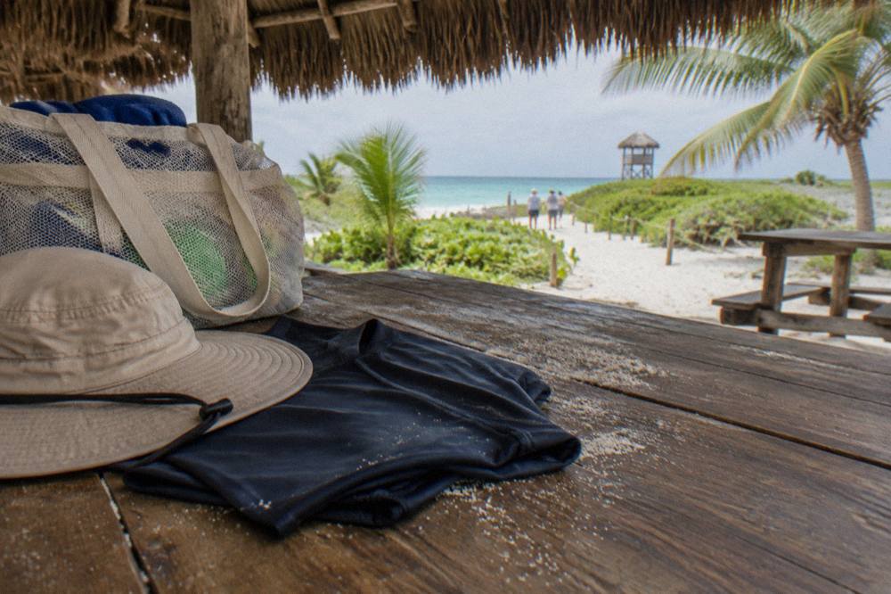 A dry rash guard and wide-brim hat folded on a wooden bench beside a mesh beach bag
